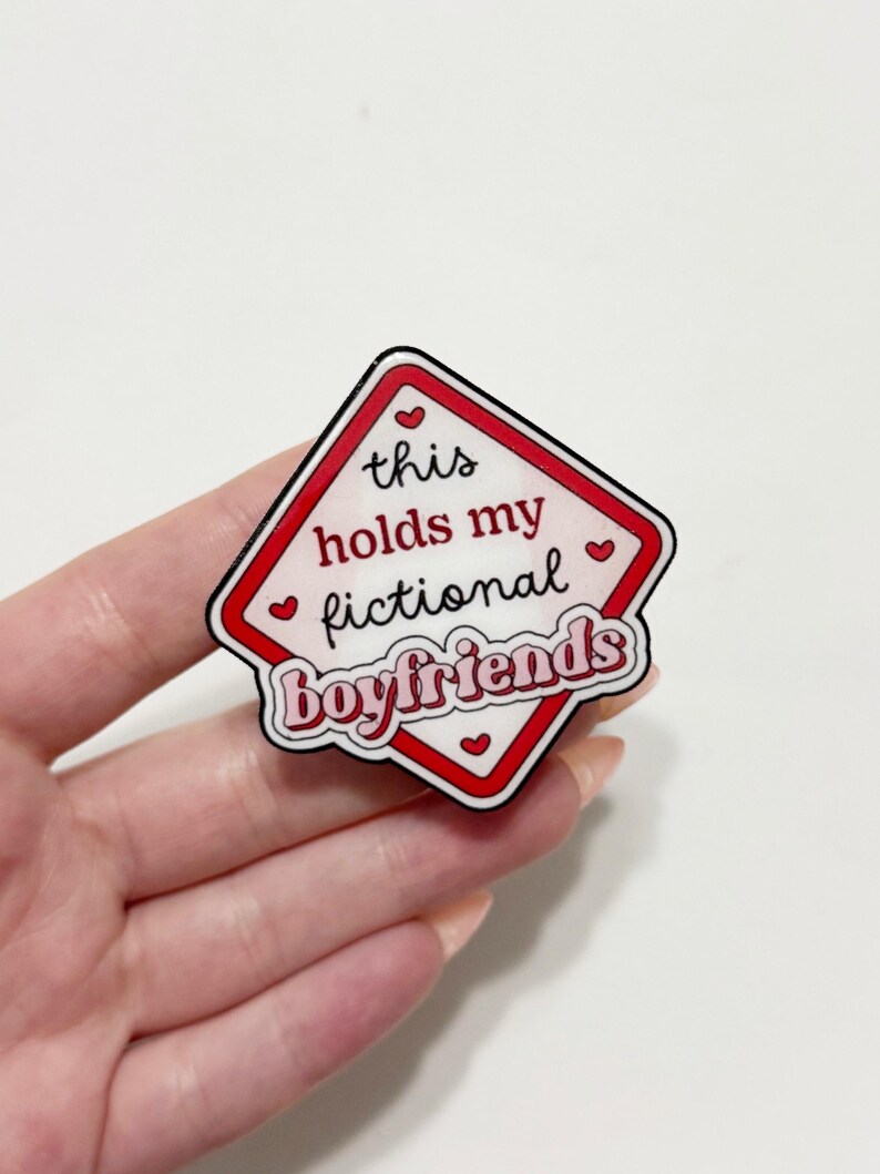 May include: A diamond-shaped pin with a red border and the text "this holds my fictional boyfriends" in a playful font. The pin has a white background with red hearts.