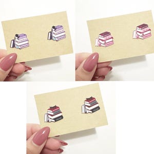 May include: A pair of book stack earrings. The earrings are made of metal and feature a stack of three books with a cat on top. The books are in shades of purple, pink, and red.