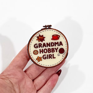 May include: A round wooden embroidery hoop with the words "GRANDMA HOBBY GIRL" and illustrations of a star, teacup, button, flower, and yarn. The design is on a cream-colored background.