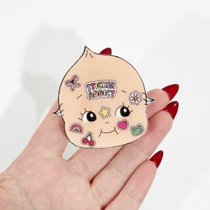 May include: A light peach-colored sticker in the shape of a head with a smiling face and various small stickers. The stickers include a bow, a flower, a star, a rainbow, a cherry, and a frog. The text "STICKER ADDICT" is also on the sticker.
