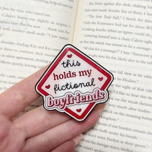 May include: A red and white diamond-shaped sticker with the text "this holds my fictional boyfriends." The sticker has a black outline and small heart accents. The sticker is placed on top of an open book.