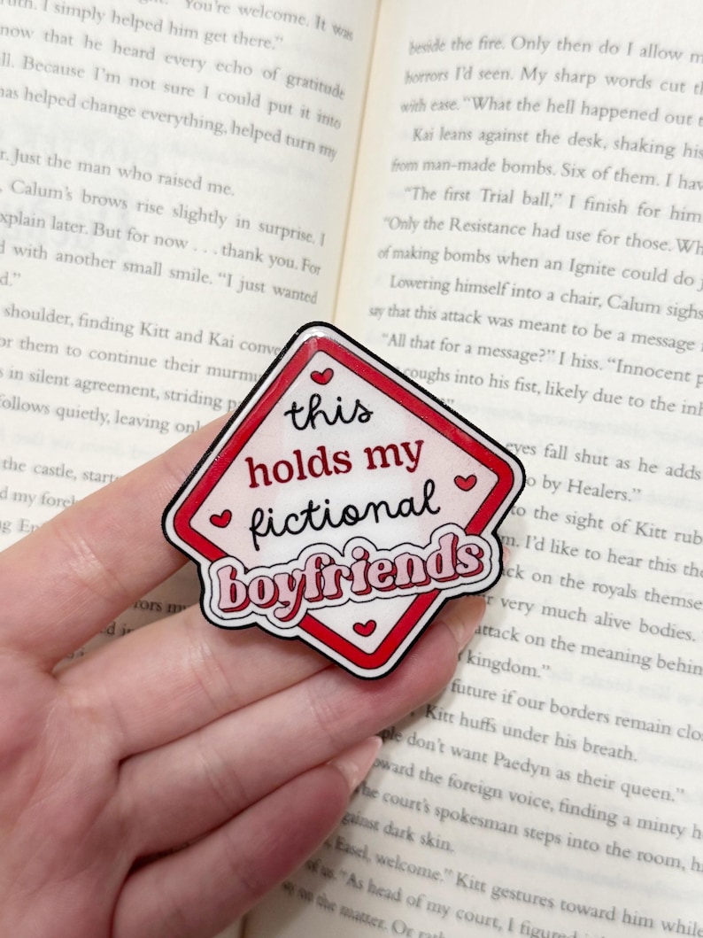 May include: A red and white diamond-shaped sticker with the text "this holds my fictional boyfriends." The sticker has a black outline and small heart accents. The sticker is placed on top of an open book.