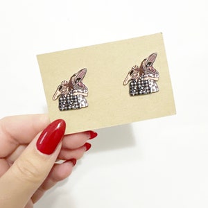 May include: A pair of novelty earrings featuring a stylized, open-mouthed skull design with a black and white checkered shirt. The earrings are displayed on a tan card, with a white background.