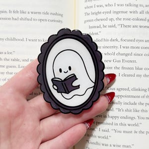 May include: A black and white sticker featuring a cartoon ghost reading a book. The ghost is inside a black oval frame with a scalloped edge. The sticker is held in a hand over an open book.