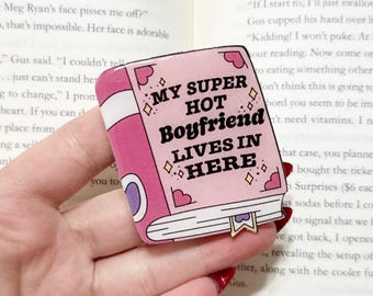 May include: A pink book-shaped pin with the text "MY SUPER HOT Boyfriend LIVES IN HERE." The pin has a white spine, a gold bookmark, and decorative hearts and diamonds. The pin is held in a hand.