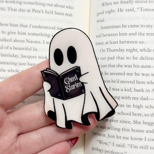 May include: A white ghost-shaped pin with black eyes and feet, holding a black book titled "Ghost Stories." The pin is a whimsical accessory, perfect for Halloween or book lovers.