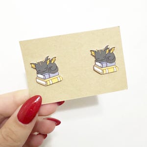 May include: A pair of black and yellow dragon book pin earrings. The dragons are curled up on top of a stack of books.