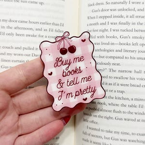 May include: A pink and white gingham-patterned bookmark with the text "Buy me books & tell me I'm pretty." The bookmark has a scalloped edge, cherries, and heart accents. It is held over an open book.
