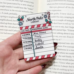 May include: A rectangular bookmark with a festive design, featuring the text "North Pole Library." The bookmark has a red and white striped border and a list of names, including Mrs. Claus and Santa.