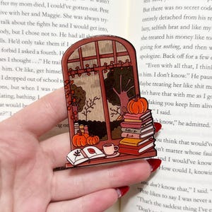 May include: A fall-themed window-shaped sticker featuring pumpkins, books, and a cup of coffee. The sticker has a warm color palette of orange, brown, and yellow, evoking a cozy autumn atmosphere. The books have titles like "Fallin' for Books".
