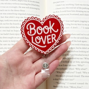 May include: A red heart-shaped badge with the words "Book Lover" in white script. The badge has a scalloped edge and a clear plastic retractable holder. The background is a book with printed text.