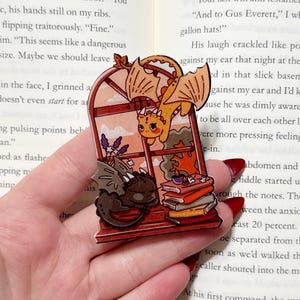 May include: A whimsical pin featuring a window scene with three cartoon dragons. One orange dragon flies above, while a brown and a black dragon rest inside the window. Books and a candle are also visible. The pin is likely made of enamel.