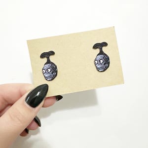 May include: A pair of black and white earrings featuring a cartoon image of a head with large eyes. The earrings are on a brown card.
