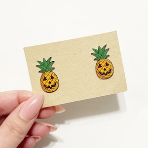 May include: A pair of wooden pineapple earrings with jack-o'-lantern faces. The earrings are yellow with black details and green tops. They are displayed on a tan card, held by a person with pink nail polish.