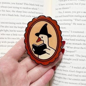May include: A sticker featuring a cartoon goose wearing a witch hat and reading a book. The goose is inside an oval frame with a scalloped edge. The sticker has a brown and orange color scheme.