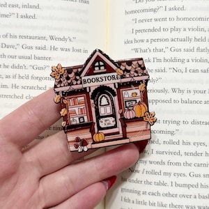 May include: A detailed bookstore-themed enamel pin. The pin features a brown roof, a door with the text "Open til late", pumpkins, and the word "BOOKSTORE". The pin is held in a hand over an open book.