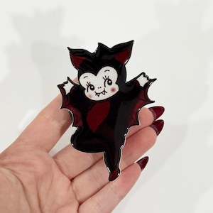 May include: A glossy enamel pin shaped like a cartoon bat with a white face, large eyes, and small fangs. The bat has black wings with dark red accents and a black body. The pin features a smooth, reflective surface with a silver-toned metal outline.