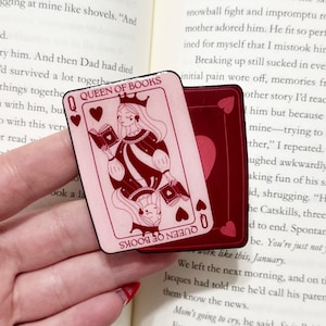 May include: A pink and burgundy Queen of Books playing card design. The card features a queen with a crown, holding a book, and the text "Queen of Books". A second card is visible behind the first.