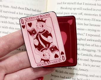 May include: A pink and burgundy Queen of Books playing card design. The card features a queen with a crown, holding a book, and the text "Queen of Books". A second card is visible behind the first.