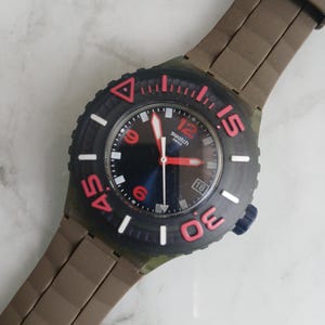 Puede incluir: Un reloj de pulsera Swatch con correa de silicona marrón oscuro y esfera negra. El reloj cuenta con un bisel negro giratorio con los números rojos 15, 30 y 45, manecillas rojas, un segundero blanco y una ventana de fecha en la posición de las cuatro.