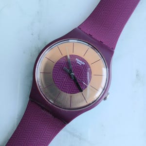 Könnte beinhalten: Eine lila Swatch-Uhr mit passendem Armband. Das Zifferblatt der Uhr ist hellorange mit schwarzen Zeigern und dem Swatch-Logo. Das Armband hat ein strukturiertes Zickzackmuster.