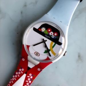Puede incluir: Reloj Swatch con esfera blanca con un sombrero negro adornado con flores de colores. El reloj tiene una correa azul claro y una banda estampada en rojo y blanco. La esfera del reloj muestra el logotipo de Swatch.