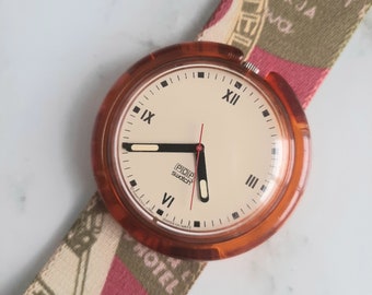 May include: A vintage orange plastic Swatch watch with a white face and black hands. The watch has a red second hand and the words "POP swatch" printed on the face. The watch is on a colorful patterned fabric strap.