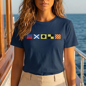 May include: Navy blue t-shirt with a nautical flag design across the chest. The shirt is a crew neck style and is paired with beige pants. The background shows a boat and the ocean.