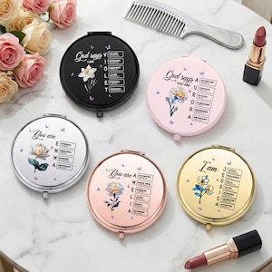 May include: A collection of round compact mirrors in various colors, including black, pink, silver, rose gold, and gold. Each mirror features floral designs, butterflies, and inspirational text. A comb and lipstick are also present.