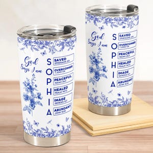 May include: Two white stainless steel tumblers with blue floral designs and the text "God Says I am." One tumbler is on a wooden surface. The text on the tumblers includes "Saved," "Overcomer," and "Abundant."