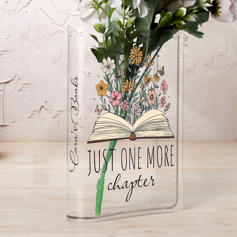 May include: Clear acrylic vase shaped like a book, with the text "Just One More Chapter." The vase is filled with flowers and butterflies. The words "Cora's Books" are written vertically on the side.
