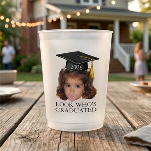 Pu&ograve; includere: Un bicchiere di plastica smerigliata con una grafica di cappello di laurea e la scritta "2026 LOOK WHO'S GRADUATED". La grafica presenta il volto di un bambino con un cappello di laurea con un nappa gialla.