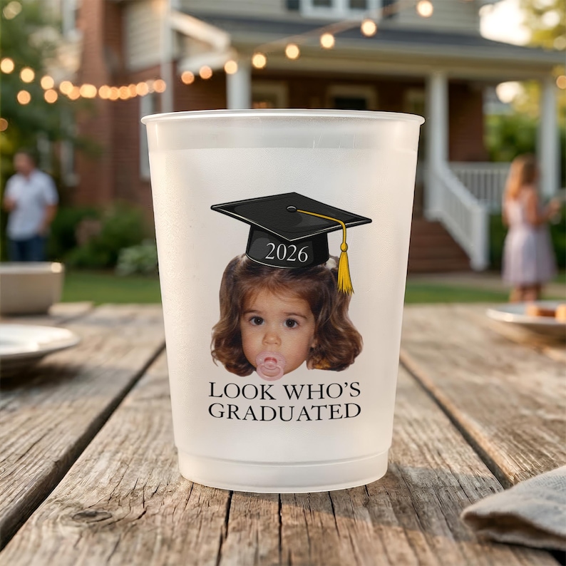 Pu&ograve; includere: Un bicchiere di plastica smerigliata con una grafica di cappello di laurea e la scritta "2026 LOOK WHO'S GRADUATED". La grafica presenta il volto di un bambino con un cappello di laurea con un nappa gialla.