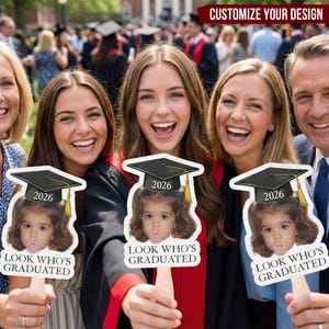 May include: A group of people smiling and holding graduation photo props. Each prop features a child's photo wearing a graduation cap with the year 2026 and the text "LOOK WHO'S GRADUATED". The background shows a graduation ceremony.
