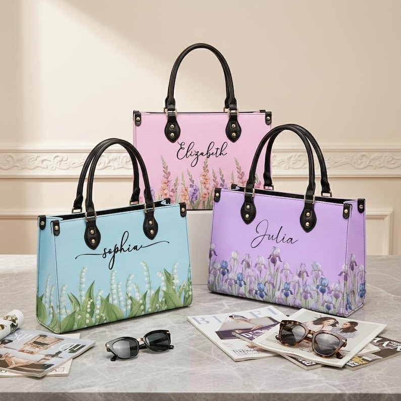 May include: Three pastel-coloured tote bags, light blue, pink, and lavender, each with a floral design and a name: Sophia, Elizabeth, and Julia. The bags have black handles and hardware. Sunglasses and magazines are in the foreground.