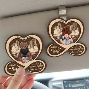 May include: Two wooden car visor ornaments with heart-shaped designs. Each features a couple embracing, with cityscapes in the background. One says "Drive Safe" and "I Fucking Love You (either way works)". The other says "Linda" and "Lucas".