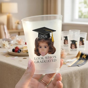Pu&ograve; includere: Un bicchiere di plastica trasparente con un'immagine stampata di un bambino con un cappello di laurea e l'anno 2026. Il testo "LOOK WHO'S GRADUATED" &egrave; stampato sotto l'immagine. Diversi bicchieri simili sono visibili sullo sfondo.