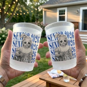 Op de afbeelding: Twee doorzichtige plastic bekers met een bedrukt ontwerp. Het ontwerp toont een zwart-wit afbeelding van een kind met een hartvormige zonnebril, de tekst "SENIOR 2026" en de naam "Sophia" in blauw. De bekers worden voor een vage buitenachtergrond gehouden.