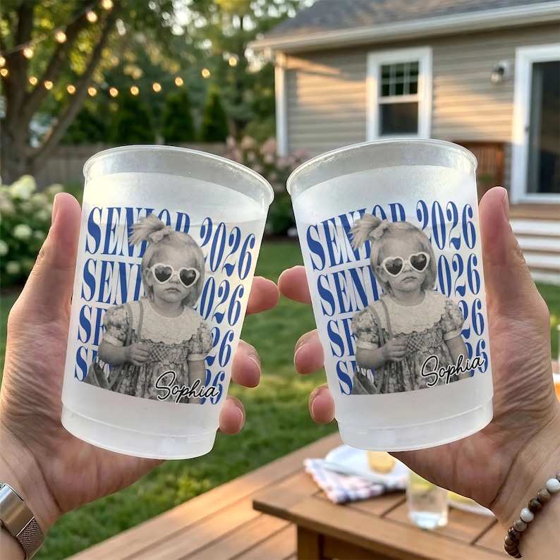 Op de afbeelding: Twee doorzichtige plastic bekers met een bedrukt ontwerp. Het ontwerp toont een zwart-wit afbeelding van een kind met een hartvormige zonnebril, de tekst "SENIOR 2026" en de naam "Sophia" in blauw. De bekers worden voor een vage buitenachtergrond gehouden.