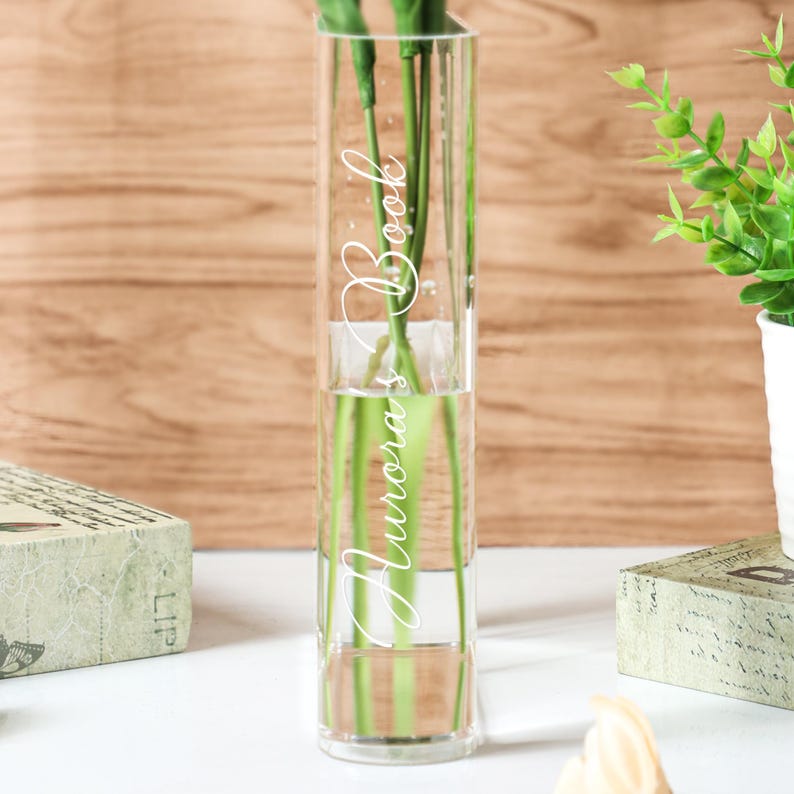 May include: A clear, rectangular glass vase with the words "Aurora's Book" in a white, cursive font. The vase contains water and green stems. The background is a light brown wooden surface.