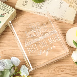 May include: A clear acrylic book cover with the text "JUST ONE MORE chapter" in white script. The cover features a floral design above an open book illustration. The cover is on a wooden surface.