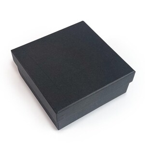 May include: A black, rectangular gift box with a textured surface. The box is closed and has a lid that fits snugly over the base. The box is sitting on a white surface.