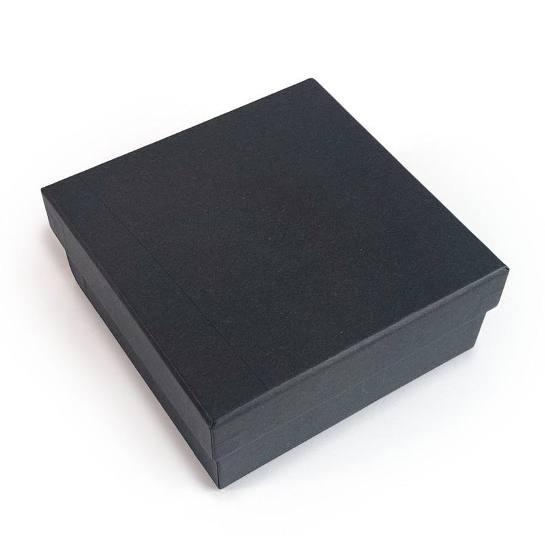 May include: A black, rectangular gift box with a textured surface. The box is closed and has a lid that fits snugly over the base. The box is sitting on a white surface.