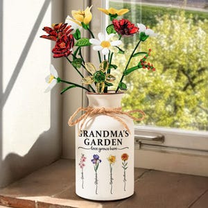 May include: A white ceramic vase with a floral arrangement. The vase is adorned with the text "GRANDMA'S GARDEN love grows here" and names below. The flowers are red, white, and yellow. A twine bow accents the vase. The vase is on a windowsill.