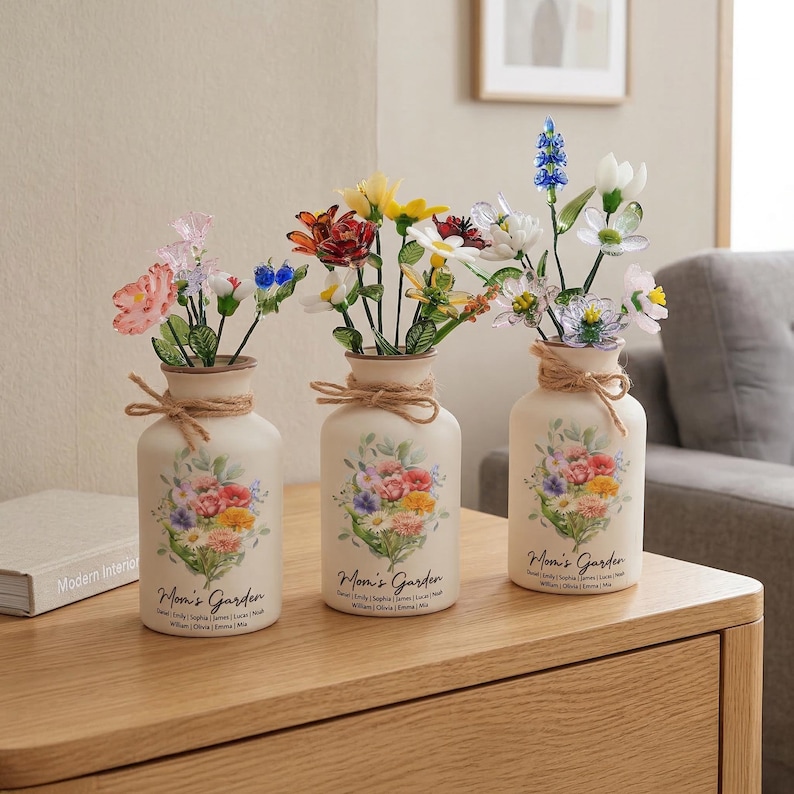 May include: Three off-white ceramic vases with floral arrangements. Each vase has a floral design and the text "Mom's Garden". The vases are tied with twine and sit on a wooden surface. The flowers are colorful and varied.