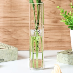 May include: A clear glass rectangular vase with the text "Cora's Books" in a cursive font. The vase contains water and green stems. The background is a light brown wooden surface. Decorative items are on either side of the vase.