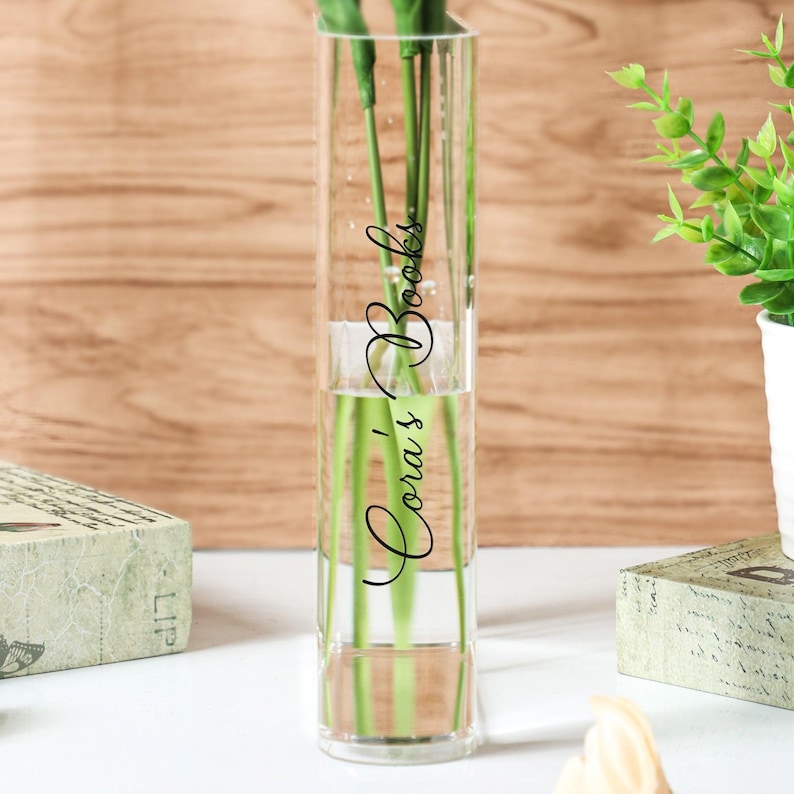 May include: A clear glass rectangular vase with the text "Cora's Books" in a cursive font. The vase contains water and green stems. The background is a light brown wooden surface. Decorative items are on either side of the vase.