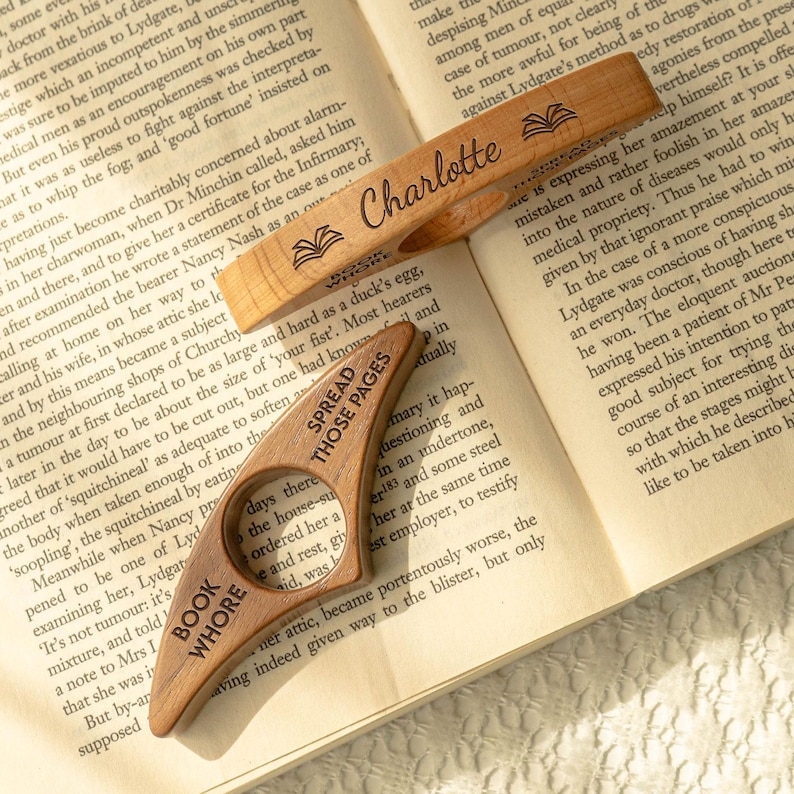 May include: Wooden book page holder with the text "BOOK WHORE" and "SPREAD THOSE PAGES". The page holder is in the shape of a finger ring and is resting on the pages of a book. The top page holder has the name "Charlotte" engraved.