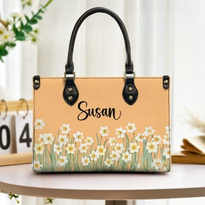 May include: Peach-coloured handbag with black handles and accents. The bag features a floral design of white daffodils with yellow centres and green stems. The name "Susan" is written in black script.