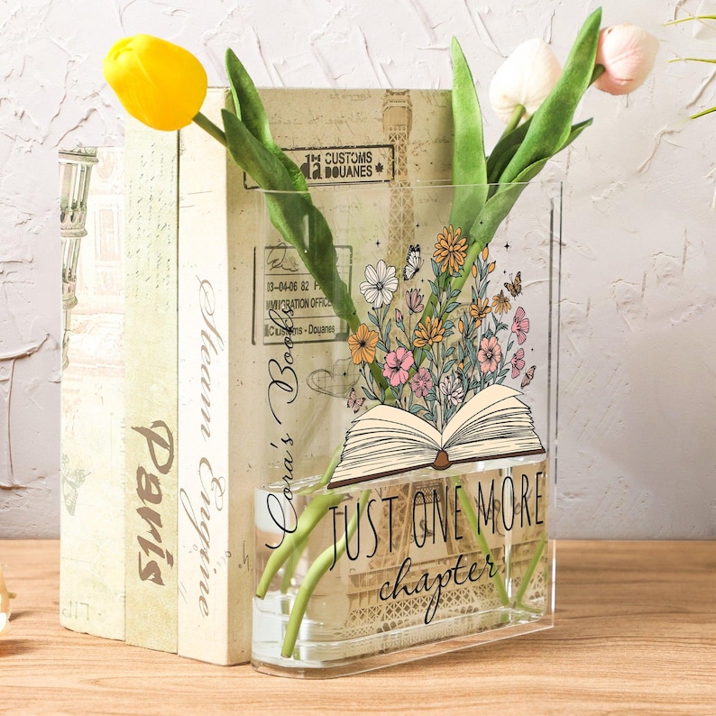 May include: A clear acrylic vase with the text "JUST ONE MORE chapter" and a floral design. The vase holds yellow and pink tulips, and is placed in front of a stack of books with the word "Paris" visible.